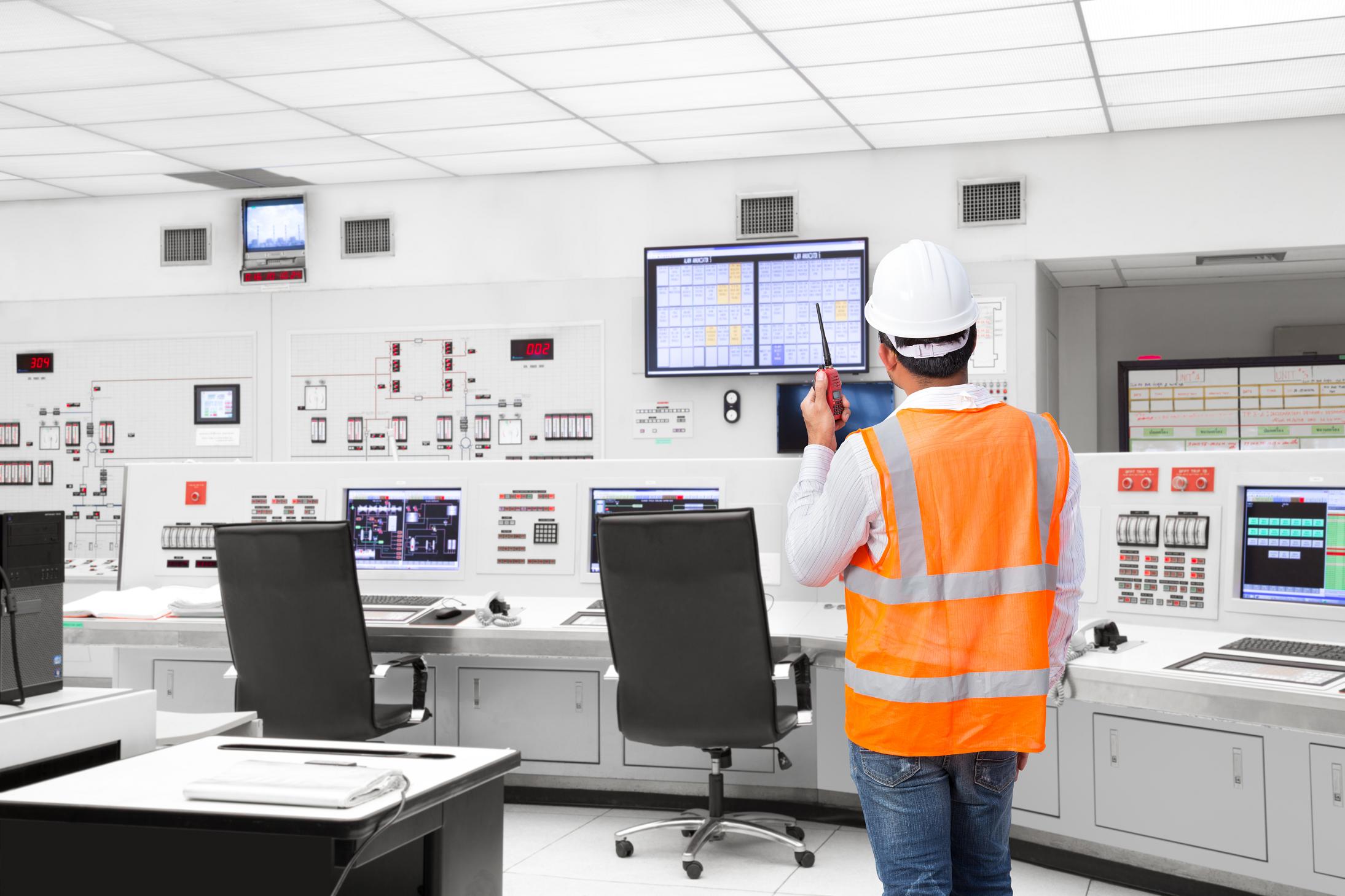 A man in a white hardhat and high-visibility vest stands in a large process plant control room holding a walkie-talkie and looking towards a screen with alarm annunciations displayed. The control room is mostly white, so the man's vest, the instruments and displays pop out of the image.